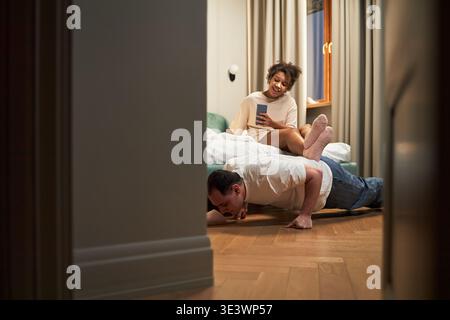Caucasian middle aged man doing push ups on wooden floor while Black young adult woman sitting on bed holding smartphone and laughing, both engaging in playful interaction in modern bedroom Stock Photo