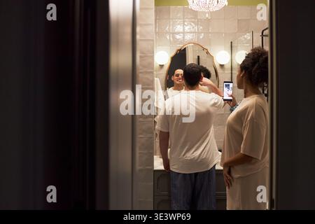 Caucasian young adult man standing in front of bathroom mirror, taking selfie with smartphone, while Black young adult woman standing nearby observing reflection in mirror Stock Photo
