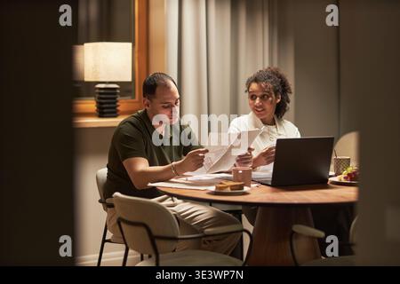 Young adult Caucasian man and young adult Black woman sitting at table reviewing documents together, using laptop, discussing paperwork, collaborating on financial or business tasks indoors Stock Photo