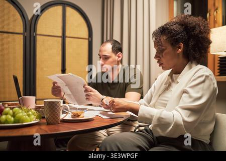 Caucasian young adult man and Black young adult woman sitting at table reviewing documents together, both focused on paperwork with laptop, coffee mugs and fruit plate visible Stock Photo