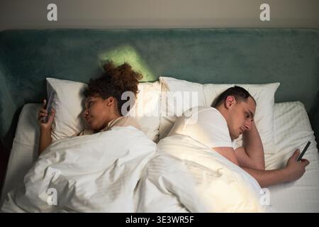 Young adult Caucasian man and young adult Black woman lying in bed side by side using smartphones, both facing away from each other, engaging with mobile devices before sleep Stock Photo