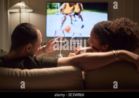 Young adult Caucasian man and young adult Black woman sitting on sofa clinking wine glasses while watching basketball game on television, relaxing together in living room Stock Photo