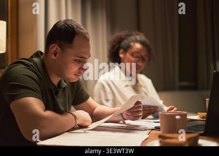 Middle aged Caucasian man reading document at desk with middle aged Black woman reviewing papers beside him, both working together in home office setting with paperwork and laptop Stock Photo