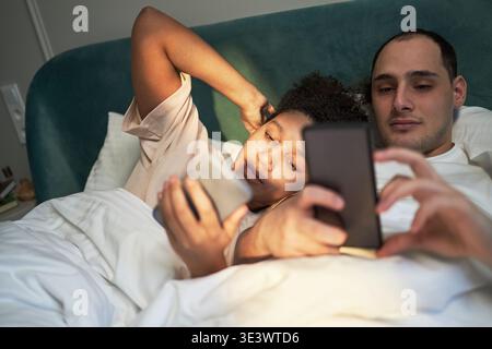 Young adult Caucasian man and young adult Black woman lying in bed using smartphones, both looking at screens with relaxed expressions, morning or evening bedroom setting Stock Photo