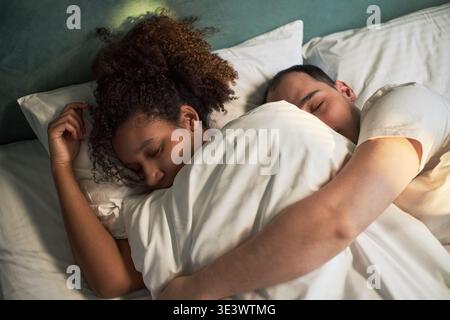 Caucasian man and Black woman sleeping together in bed, embracing under blanket, both appearing as young adults, peaceful expressions, heads resting on pillows, eyes closed Stock Photo