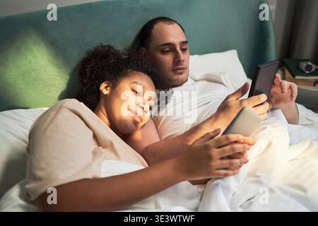 Young adult Black woman and young adult Caucasian man lying in bed using smartphone and tablet, both focused on screens, relaxing together in bedroom during leisure time Stock Photo