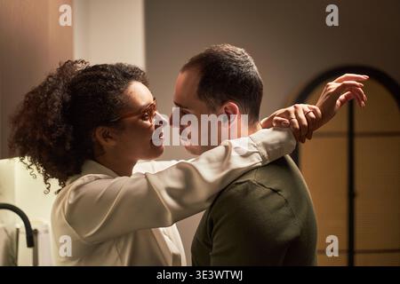 Young adult Black woman embracing young adult Caucasian man in intimate moment, standing close together in modern indoor setting, both gazing into each others eyes, showing affection Stock Photo