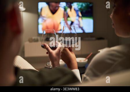 Young adult Caucasian man and young adult Black woman clinking wine glasses while watching television together, sitting close on couch with television screen visible in background Stock Photo