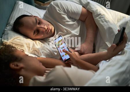 Young adult Caucasian man lying in bed looking at young adult Black woman using smartphone for online grocery shopping, both relaxing under blanket in bedroom at night Stock Photo