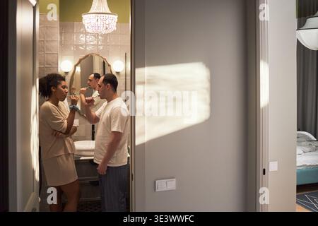 Multiethnic group of young adult men and woman brushing teeth together in bathroom, standing near sink and mirror, engaging in morning hygiene routine, bedroom visible in background Stock Photo