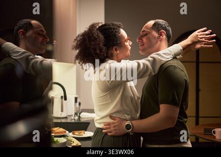 Young adult Black woman embracing young adult Caucasian man in kitchen, both standing close and gazing at each other, reflection visible in background, food and drink on counter Stock Photo