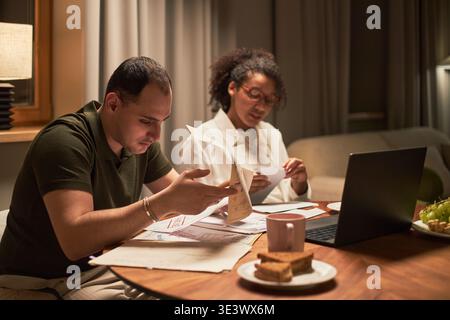 Middle aged Caucasian man and middle aged Black woman sitting at table reviewing financial documents together, using laptop, holding papers, coffee mug and snacks on table Stock Photo