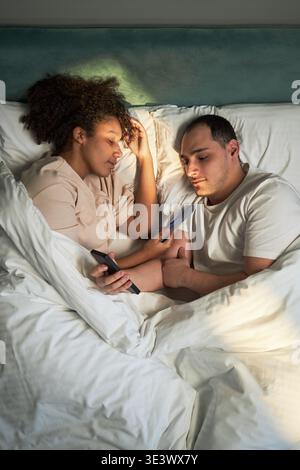 Vertical of Young adult Black woman and young adult Caucasian man lying in bed using smartphones, holding hands while looking at screens, relaxing together in bedroom setting Stock Photo