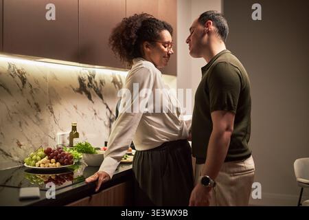Middle aged Black woman and middle aged Caucasian man standing close together in modern kitchen, woman leaning against counter with fresh fruits and vegetables, couple sharing intimate moment Stock Photo