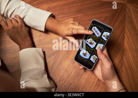 Caucasian young man and Black woman browsing food delivery app on smartphone, hands of couple visible on wooden table, one woman pointing at menu images on screen Stock Photo