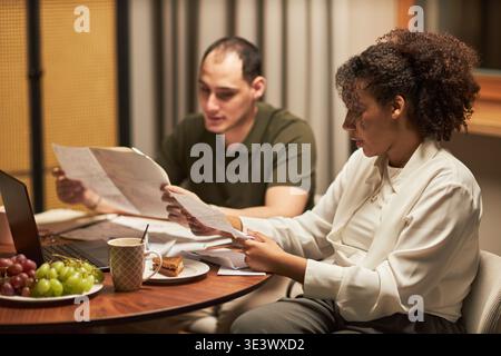 Caucasian young adult man and Black young adult woman sitting at table reviewing documents together, working with laptop and papers, coffee mug and fruit plate visible on table Stock Photo
