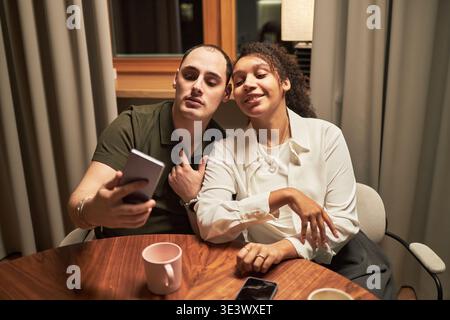 Young adult Caucasian man and young adult Black woman sitting at table taking selfie together using smartphone, smiling and posing closely, coffee mugs and smartphone on table Stock Photo