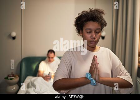 Young adult Black woman meditating with eyes closed and hands in prayer position in foreground, Caucasian middle aged man sitting on bed in background using smartphone Stock Photo
