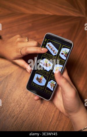 Vertical of Caucasian young adult woman holding smartphone while Black young adult woman pointing at food photos on screen, selecting meal for delivery from digital menu, wooden table background Stock Photo