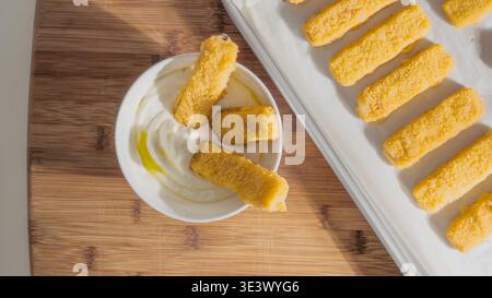 A bowl of creamy white sauce sits next to a tray of breaded fish sticks on a wooden board. Stock Photo