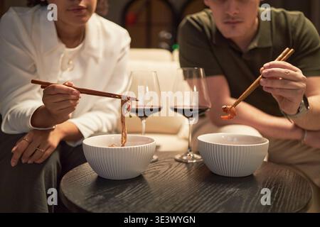 Young adult Black woman and young adult Caucasian man sitting together eating noodles with chopsticks from bowls, drinking red wine, focusing on food in casual indoor setting Stock Photo