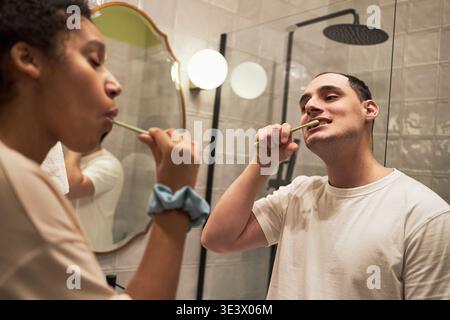 Caucasian young adult man and Black young adult woman brushing teeth together in modern bathroom, standing side by side in front of mirror, focusing on dental hygiene routine Stock Photo