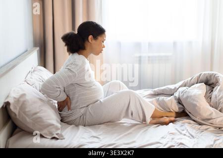 Pregnant Black woman sitting on bed holding her lower back in pain Stock Photo