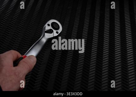 Close up of hand holding ratchet wrench over dark textured background. Mechanical tool ready for use in workshop or repair setting with copy space. Stock Photo
