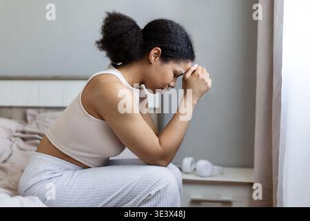 Upset black woman sitting on bed with hands clasped and head down Stock Photo