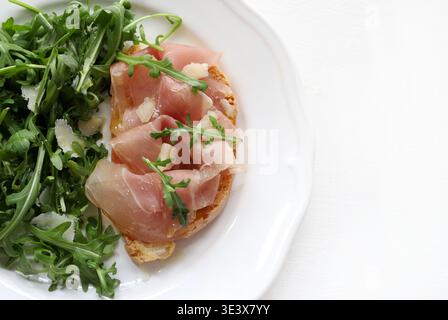 Bruschetta with prosciutto, fresh arugula, and parmesan shavings on a white background. A typical Italian dish. Stock Photo