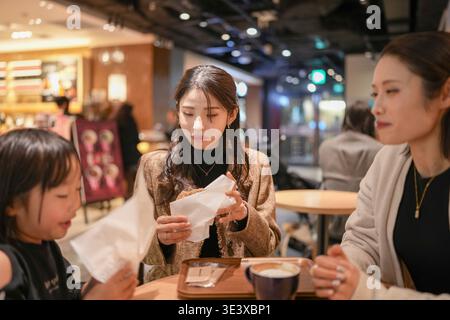 Three women sit at a cafe table, engaged in quiet conversation. One holds a napkin, eyes focused on something unseen. Another leans forward, listening Stock Photo