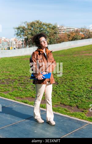 Professional woman with glasses and a brown leather jacket standing in a city park, holding a blue folder, looking away from the camera on a sunny day Stock Photo