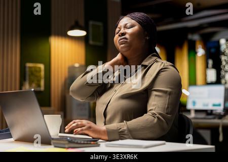 Female assistant in discomfort due to neck stiffness and backache, doing self massage to relieve the pain and strain. Businesswoman struggling with incorrect posture at the office. Stock Photo