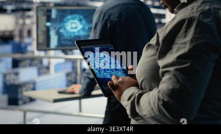 Close up of data center system administrators deploying deep learning software on rigs. Team of server hub IT experts train AI systems with datasets using tablet to support applications, camera B Stock Photo