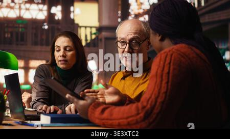 Mature academic colleagues prepare documents for university coursework or exams, transcribing essential ideas from textbooks. Research and education in a quiet public library. Stock Photo