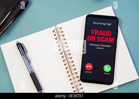 Smartphone on desk displays incoming scam call alert next to notebook and pen, illustrating fraud attempt, cyber security risk, workplace awareness, d Stock Photo