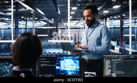 Manager supervising work of server room engineer using AI to review infrastructure diagnostics on devices. Tech company supervisor overseeing IT expert in data center analyzing data using AI, camera A Stock Photo