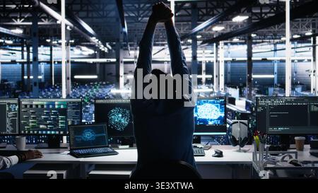 Server room technician stretching sore back while using AI at desk. Fatigued IT expert in data center relaxing strained muscles, feeling backache while using artificial intelligence, camera A Stock Photo