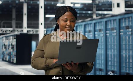 Programmer in data center using laptop to upgrade systems for increased stability. IT professional in server farm configuring infrastructure with notebook for optimum performance, camera B Stock Photo