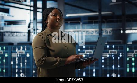 Admin in data center using app on laptop to ensure error free rigs networks for intense computational demands. IT expert uses notebook to monitor server hub gear components for failures, camera B Stock Photo