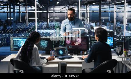 Data center manager coordinating programmers fixing critical error while using AI. Server room team leader overseeing workers repairing glitch popup while using artificial intelligence, camera A Stock Photo