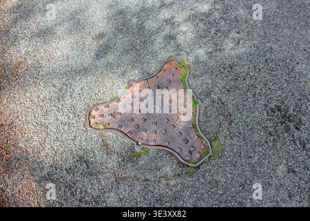 An aged, rusted triangular manhole cover with rounded edges is embedded in a weathered asphalt surface, featuring embossed text and square patterns. Stock Photo