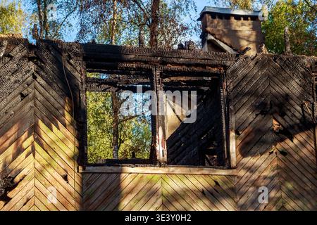 Charred remnants of a wooden house after a fire, showcasing burned walls and an empty window frame under a clear autumn sky, reflecting tragedy and de Stock Photo