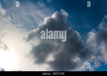 Dramatic, dark storm clouds gathering in a vibrant blue sky, with bright sunlight peeking through lighter wispy formations. A moody, evolving weather Stock Photo