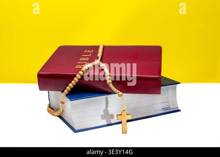 Two stacked Bibles with a wooden rosary and cross on a bright yellow and white background. Represents Christian faith, prayer, devotion, and modern re Stock Photo