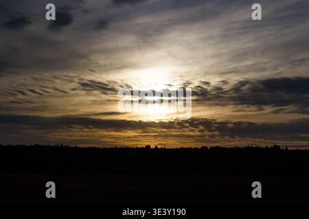 Intense sunset peeks through stormy clouds above a dark forest line. Golden light bursts across the vast, moody sky at dusk Stock Photo