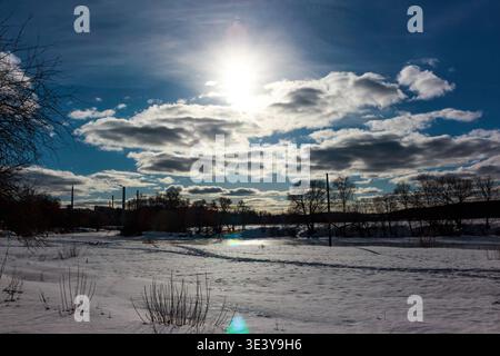 Sunlight pierces through dramatic clouds, illuminating a vast snow-covered landscape with scattered trees and distant industrial structures, a true wi Stock Photo