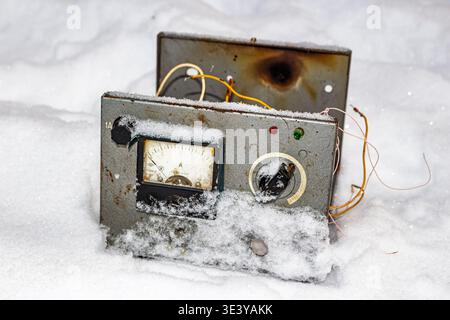 Weathered Soviet-era electronic device, an ammeter and wires exposed, half-buried in fresh winter snow. A defunct relic of industrial tech, forgotten Stock Photo