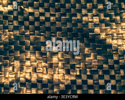 Golden light dances across a woven carbon fiber surface. Dark and bright squares interlock in a repeating grid. The texture is rigid, yet softly refle Stock Photo