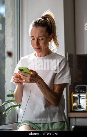 Smiling relaxed woman using scrolling smartphone standing in kitchen near window, calm start of day. Stock Photo